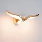 Sur un fond blanc on voit une application murale en forme d'oiseau dorée allumée.