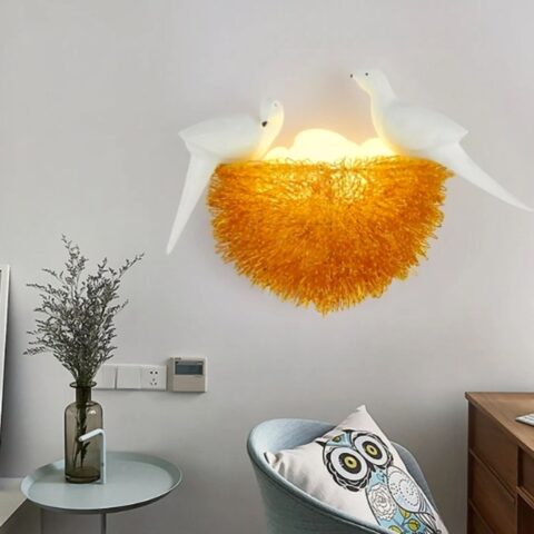 Au milieu de l'image on voit une application murale en forme de nid d'oiseau. En bas à gauche de l'image on voit une table avec un vase gris transparent avec des branchages dedans. Dans le coin en bas à droite on voit le haut d'un fauteuil avec un coussin à motif hibou dessus.