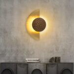 Sur un mur gris foncé on voit une applique murale au centre accrochée en forme de demi-lune dorée et allumée.