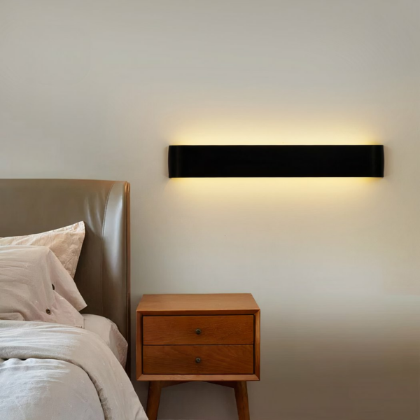Applique murale rectangle noire, avec lumière intégré. Accroché à un mur blanc à côté d'un lit, au-dessus d'une table de nuit en bois.