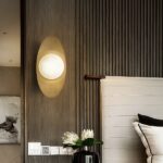Applique murale en cuivre élégante, couleur dorée avec une boule dessus qui envoi la lumière, à côté d'un lit dans une chambre