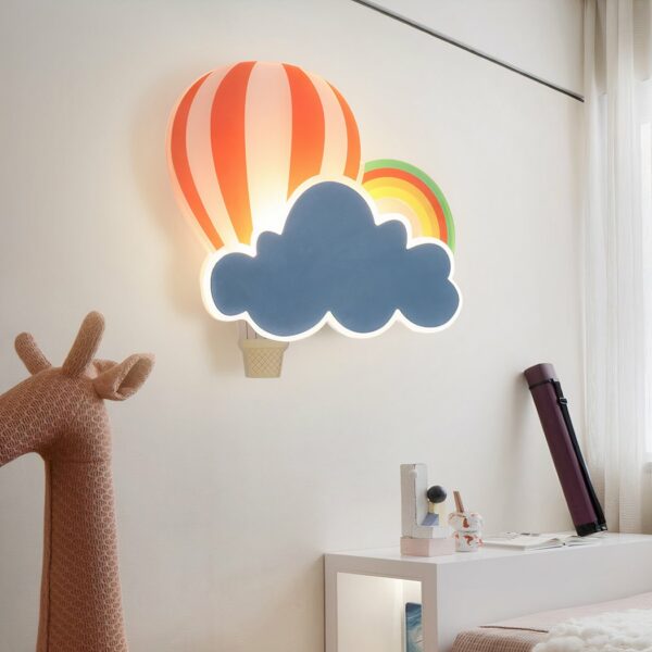 Sur le mur d'une chambre pour enfant au voit au centre de l'image une applique murale en forme de nuage, montgolfière et arc en ciel pour chambre d'enfant. en bas à gauche de l'image il y a une tête de jouet girafe rose.