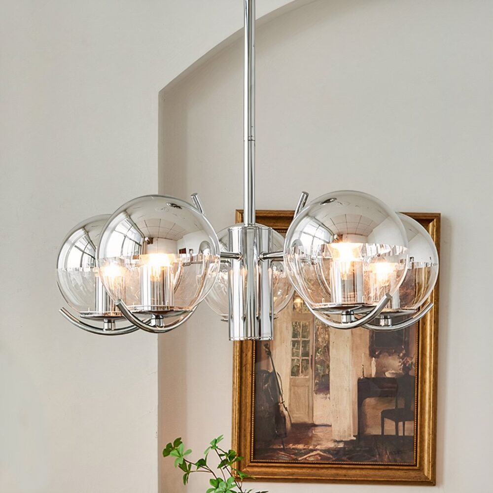 Lustre en métal argenté, avec 5 boules en verre, disposée en cercle autour d'un axe central. Installé dans une pièce aux murs beige, on voit le bout d'un rideau blanc sur la gauche et une table avec un livre et un vase en dessous du lustre, ainsi qu'un bout de tableau sur la droite.