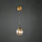 Applique murale moderne, qui se compose d'un socle rond doré, duquel part une tige fine dorée vers le bas, au bout de laquelle se trouve une boule en verre avec une ampoule à l'intérieur allumé. Sur un mur noir.