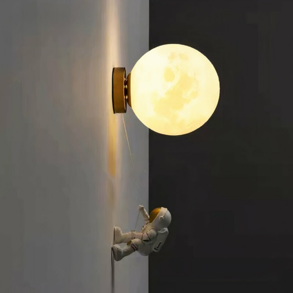 Applique murale en forme de boule accrochée au mur qui représente la lune, à laquelle est accrochée une corde. Au bout de cette corde, une figurine d'astronaute qui s'appuie contre le mur pour monter jusqu'à la lune. La lumière est allumée, installé sur un mur blanc dans une pièce dans le noir.