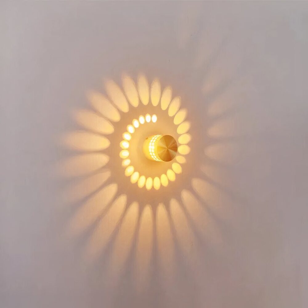 Applique murale a effet, se compose d'un cylindre métalique gris, perferé de petit trous, qui laisse diffusé la lumière en forme de spirale au mur. Allumée. Accroché sur un mur blanc.