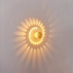Applique murale a effet, se compose d'un cylindre métalique gris, perferé de petit trous, qui laisse diffusé la lumière en forme de spirale au mur. Allumée. Accroché sur un mur blanc.
