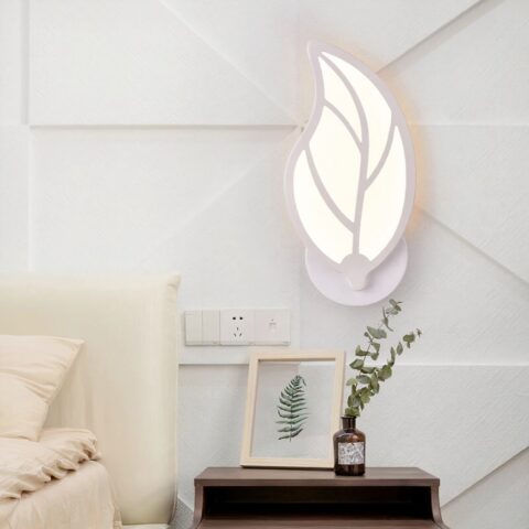 Applique murale blanche en forme de feuille plaquée au mur, allumée. Elle est accrochée sur un mur blanc près d'un lit, au-dessus d'une table de chevet e bois marron foncé. Avec une petite plante verte dessus.