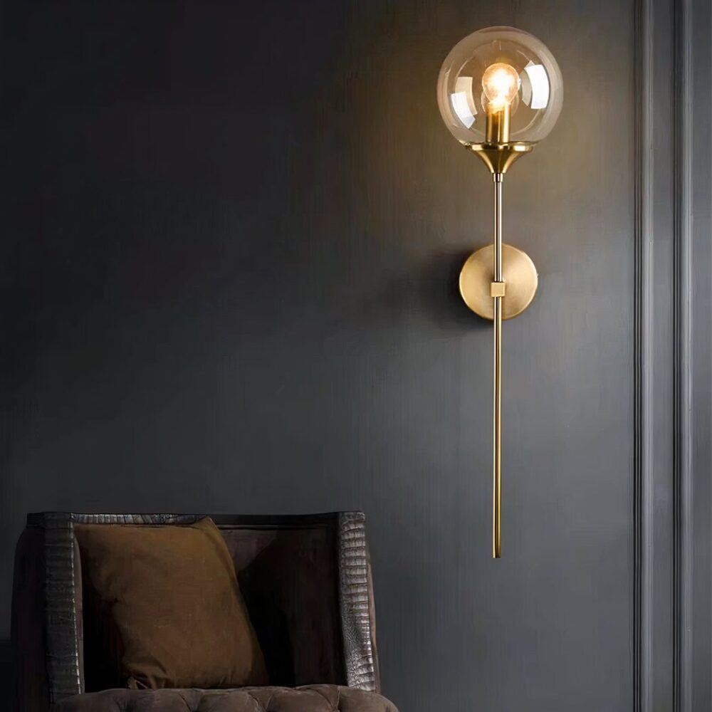 Applique murale moderne et épuré, se compose d'une tige fine dorée, sur laquelle se trouve une boule en verre avec une ampoule à l'intérieur allumé. La tige est fixée au mur par un socle rond doré aussi. Sur un mur noir. À côté se trouve un fauteuil, dont on ne voit que le dossier marron.