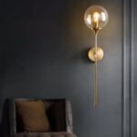Applique murale moderne et épuré, se compose d'une tige fine dorée, sur laquelle se trouve une boule en verre avec une ampoule à l'intérieur allumé. La tige est fixée au mur par un socle rond doré aussi. Sur un mur noir. À côté se trouve un fauteuil, dont on ne voit que le dossier marron.