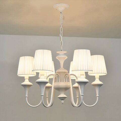 Lustre blanc style baroque, en forme de chandelier avec 6 abat-jours blanc, les ampoules sont allumées. Suspendu par une chaine à un plafond blanc, on voit un bout de mur blanc dans le fond.