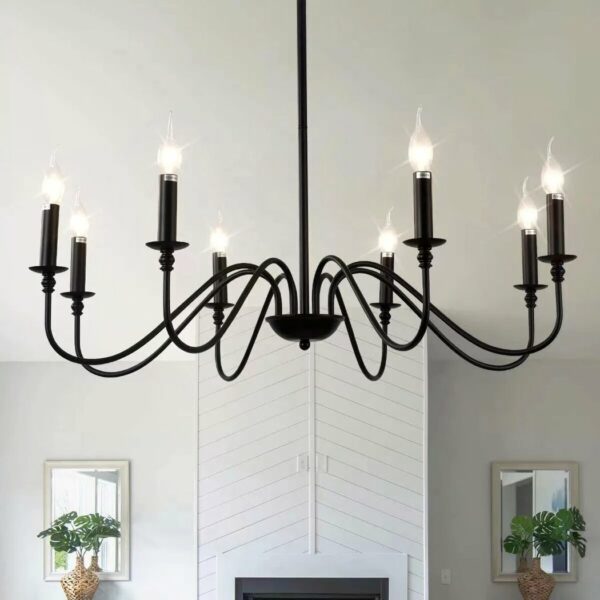 Lustre noir en fer en forme de chandelier, avec 8 bougies à ampoule. Simple et épuré. Installé dans une pièce de vie. On voit au fond une cheminé blanche avec un miroir et une plante devant le miroir de chaque côté de la cheminé.