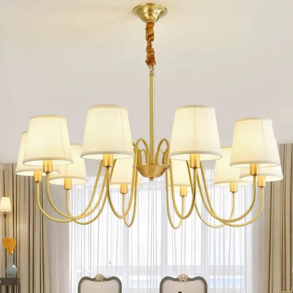 Lustre doré en forme de chandelier avec 10 têtes avec abat-jours blanc, suspendu par une chaine. On voit une fenêtre dans le fond et le dossier de deux chaises.