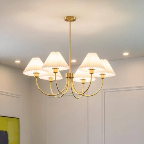 Lustre doré avec corps en cuivre et 6 abat-jours en tissu blanc, en accordéon. Accroché à un plafond blanc, on voit 3 spots lumineux dans le fond et un mur blanc avec des moulures.
