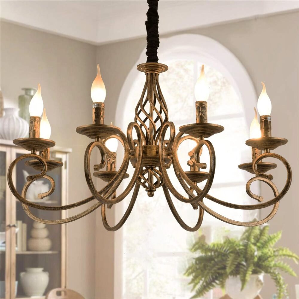 Lustre en laiton doré style chandelier avec 6 têtes avec des ampoules qui remplacent les flemmes des bougies. Les ampoules sont allumées. Le lustre est suspendu au plafond d'un salon, on voit sur la droite une plante verte à côté d'une fenêtre, et sur la gauche un morceau de meuble ouvert avec des bibelots, de la vaisselle. Les murs sont beiges.