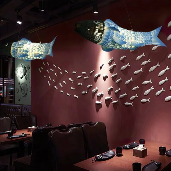 Salle de restaurant avec deux lustres en forme de gros poisson au corps bleu et blanc et la tête en baguettes de bois noir. On voit des tables dressées et un mur rouge en arrière-plan avec pleins de poissons accroché au mur comme un banc de poissons dans la mer.