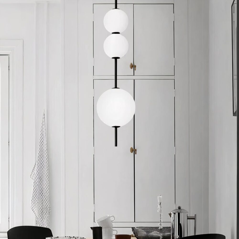 Lustre suspendu à la verticale composé de trois boules blanches allumées glissées sur un axe. La boule de bas est plus grande que les deux petites en haut. Dans le fond, on voit un mur blanc et des portes de placard blanc.