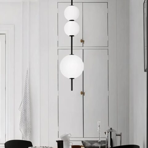 Lustre suspendu à la verticale composé de trois boules blanches allumées glissées sur un axe. La boule de bas est plus grande que les deux petites en haut. Dans le fond, on voit un mur blanc et des portes de placard blanc.