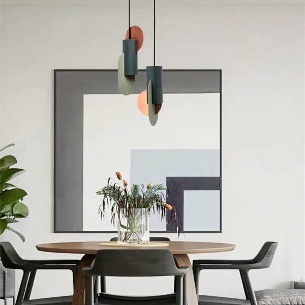 Lustres design art-déco moderne, suspendus au-dessus d'une table ronde avec un vase avec des fleurs. Il y à trois chaises autour de la table, dans le fond un mur blanc et un grand tableau avec du gris, blanc, bleu et noir en forme de L.