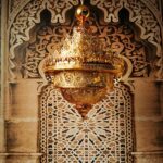 Grand lustre doré marocain, majestueux, en haut plusieurs couronnes qui donnent l'effet des flemmes, le bas descend en pointe. Tout le lustre est évidé et ajouré. Derrière un mur typique marocain avec une partie en bois sculpté et l'autre des pierres et des carreaux aux motifs marocains.