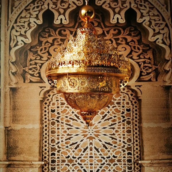 Grand lustre doré marocain, majestueux, en haut plusieurs couronnes qui donnent l'effet des flemmes, le bas descend en pointe. Tout le lustre est évidé et ajouré. Derrière un mur typique marocain avec une partie en bois sculpté et l'autre des pierres et des carreaux aux motifs marocains.