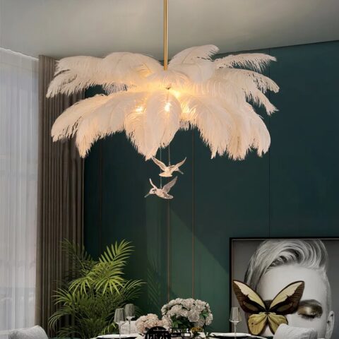 Lustre avec grandes plumes blanches, et deux figurines transparentes d'oiseaux. Suspendus. Les ampoules sont allumées et camouflées dans les plumes. On voit un mur vert foncé dans le fond, ainsi qu'un tableau et une plante verte.