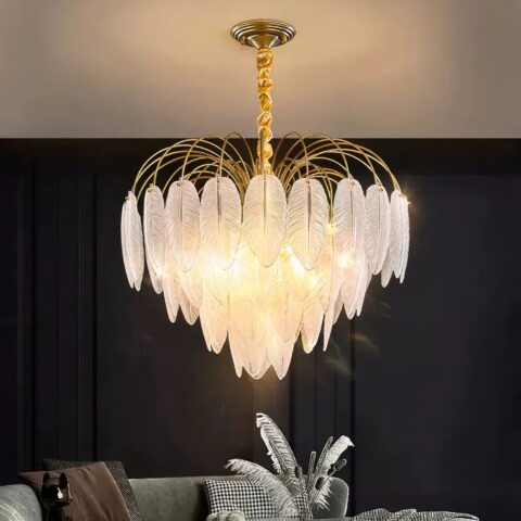 Lustre plumes en verres en forme de grappe. Allumé et installé dans un salon avec le mur du fond noir et un canapé gris. Le plafond est blanc et on voit le reflet de la lumière dessus.