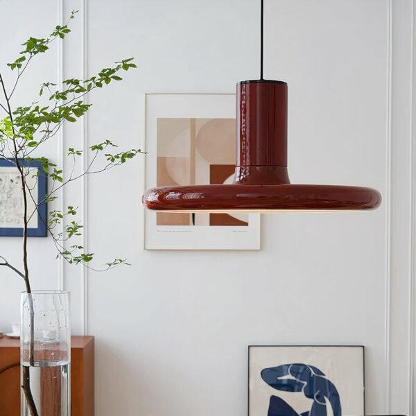 Lustre rouge rond, plat vintage, suspendu. Au fond mur blanc avec deux tableaux et à gauche un vase haut avec de l'eau et une plante verte.
