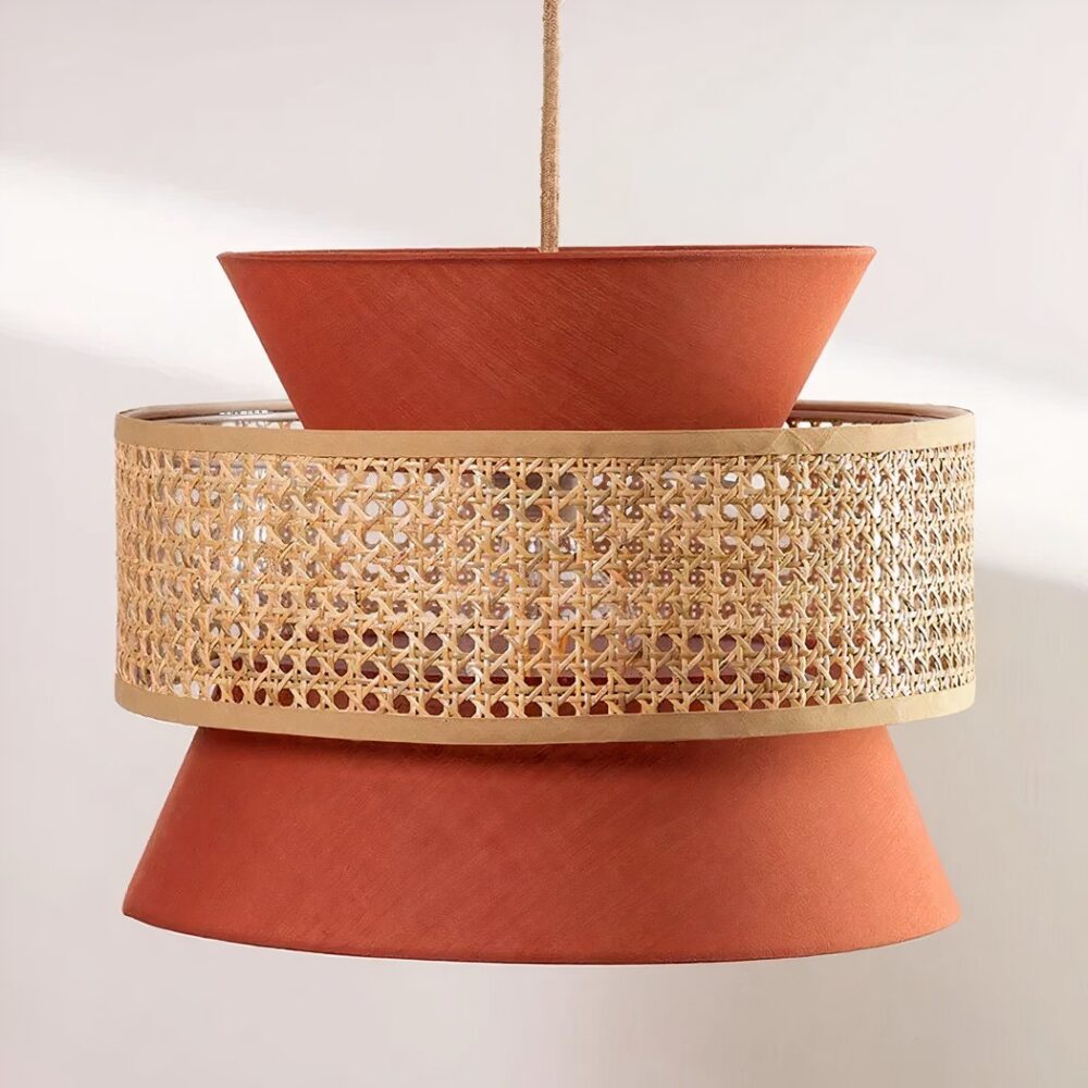 Lustre terracotta et beige, suspendu par un câble au centre. Composé d'un abat-jour en tissu couleur terracotta en forme de sablier, et d'un cercle en cannage qui entoure le sablier au centre. Sur un fond gris très clair.