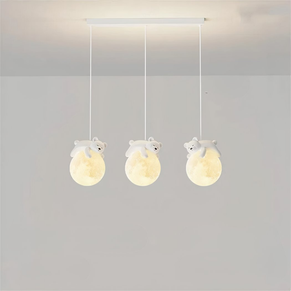 Luminaire suspendus, composé de trois câbles avec chacun une boule qui représente la lune, et un nounours qui dort sur la lune. Les boules sont allumées. Le socle rectangulaire est accroché au plafond gris clair, on voit dans le fond un mur gris clair aussi.