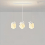 Luminaire suspendus, composé de trois câbles avec chacun une boule qui représente la lune, et un nounours qui dort sur la lune. Les boules sont allumées. Le socle rectangulaire est accroché au plafond gris clair, on voit dans le fond un mur gris clair aussi.