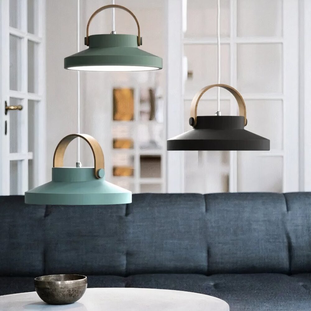 Trois suspensions en forme de macaron, avec une anse en bois suspendues au-dessus d'un canapé gris et d'une table ronde blanche. En bas à gauche, une bleue clair, au-dessus d'elle décalé sur la gauche une vert olive, et sur la droite une noir profond. On voit dans le fond une porte-fenêtre blanche à carreaux.