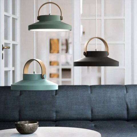 Trois suspensions en forme de macaron, avec une anse en bois suspendues au-dessus d'un canapé gris et d'une table ronde blanche. En bas à gauche, une bleue clair, au-dessus d'elle décalé sur la gauche une vert olive, et sur la droite une noir profond. On voit dans le fond une porte-fenêtre blanche à carreaux.