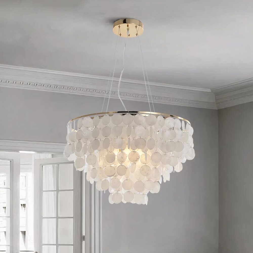 Suspension ronde avec pleins de perles de nacre ronde et plates suspendues en cascade en forme conique. On voit un plafond gris clair, et le mur du fond gris clair avec une porte-fenêtre blanche à carreaux.