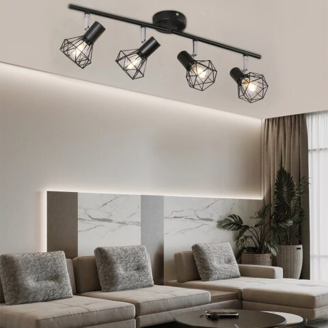 Lustre plafonnier noir de style industriel avec 4 ampoules de style spots industriels alignés. Dans un salon avec grand canapé d'angle beige avec 3 coussins rectangulaires. Une plante verte dans l'angle à droite du canapé et un rideau marron derrière elle.