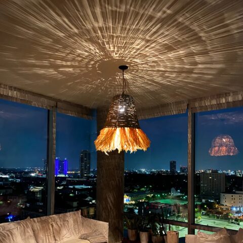 Lustre paille et raphia tissé main, accroché au plafond et allumé, dans une pièce de vie type salon. Illumine le plafond de ses jeux d'ombres grâce au tissage du raphia et de la paille.