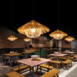 Lustre paille naturelle style paillote exotique accrochée au mur et allumée, au dessus de tables de ce qui semble etre un restaurant et dans une ambiance de lumière tamisée.
