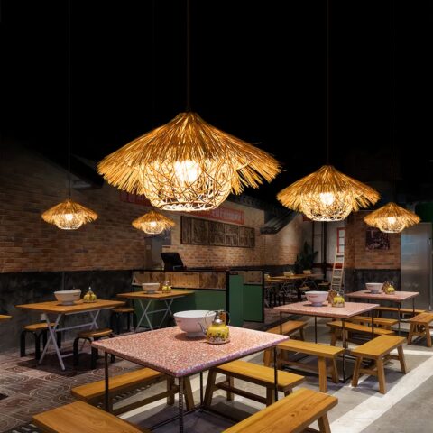 Lustre paille naturelle style paillote exotique accrochée au mur et allumée, au dessus de tables de ce qui semble etre un restaurant et dans une ambiance de lumière tamisée.
