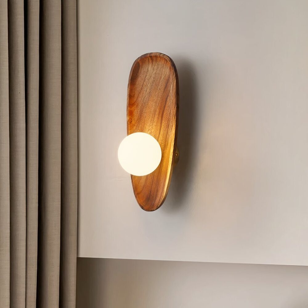 Applique murale en bois style coquillage avec sa perle lumineuse grand modèle, accrochée sur un mur blanc, à côté d'un rideau couleur taupe.