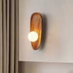 Applique murale en bois style coquillage avec sa perle lumineuse grand modèle, accrochée sur un mur blanc, à côté d'un rideau couleur taupe.