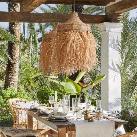 Lustre en paille artisanal tressé à la main suspendu sous une pergola et au-dessus d'une table à manger au milieu d'arbres luxuriants.