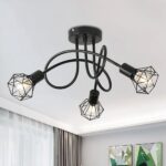 Lustre plafonnier avec 3 tiges noires entremêlées avec aux extrémités une ampoules allumées avec une grille style industrielle qui l'entoure. On aperçoit au mur en face 2 cadres, et sur la gauche une fenêtre avec rideau glissant marron.