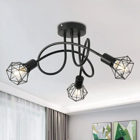 Lustre plafonnier avec 3 tiges noires entremêlées avec aux extrémités une ampoules allumées avec une grille style industrielle qui l'entoure. On aperçoit au mur en face 2 cadres, et sur la gauche une fenêtre avec rideau glissant marron.