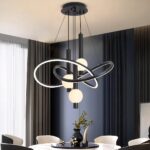 Lustre noir avec support noir fixé au plafond d'où descendent des câbles tenant des structures arrondies entrelacées lumineuses et noires à l'extérieur. Au centre de ces structures, sont pendus des tubes noirs avec une boule lumineuse allumée. Fixé au-dessus d'une table blanche ronde style marbre, avec plateau noir rond, un vase dessus et des objets de décoration. 6 fauteuils autour de la table,à gauche une baie vitrée avec un rideau gris et le mur en face est bleu canard avec des rainures verticales.