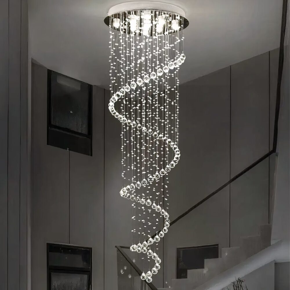 Lustre plafonnier en forme de spirale de suspensions en gouttes d'eau. Lumière blanche allumée par plusieurs LED dans le support rond argenté fixé au plafond. Derrière, on voit un escalier avec rambarde noire et des tableaux sur le mur.