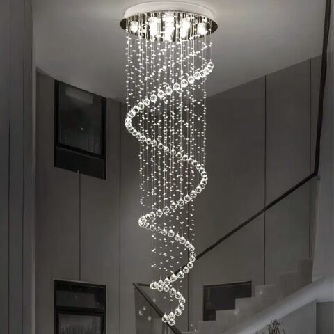 Lustre plafonnier en forme de spirale de suspensions en gouttes d'eau. Lumière blanche allumée par plusieurs LED dans le support rond argenté fixé au plafond. Derrière, on voit un escalier avec rambarde noire et des tableaux sur le mur.