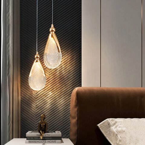 Lustre suspendu avec 2 gouttes en acrylique tenues par une structure en métal doré et un fil fin. Allumées et suspendues au-dessus d'une table de chevet avec un bouddha, à droite une tête de lit marron, et un coussin blanc.