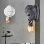Deux appliques murales rondes avec la tête d'un gorille en 3D tenant dans sa bouche 3 cordes avec une ampoule suspendue à chacune. Sur le mur gris en face, l'applique est blanche, sur le mur gris de droite, elle est noire. Sous la blanche, une table ronde noire avec un seul pied fin. Dessus un vase transparent avec un feuillage, des livres et une tasse sur la pile de livres.