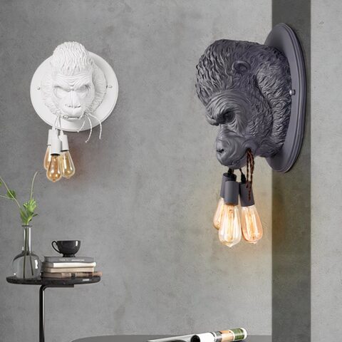 Deux appliques murales rondes avec la tête d'un gorille en 3D tenant dans sa bouche 3 cordes avec une ampoule suspendue à chacune. Sur le mur gris en face, l'applique est blanche, sur le mur gris de droite, elle est noire. Sous la blanche, une table ronde noire avec un seul pied fin. Dessus un vase transparent avec un feuillage, des livres et une tasse sur la pile de livres.