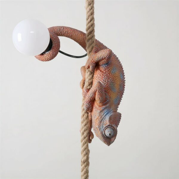 Suspension d'un caméléon marron avec du bleu par endroit, tête vers le bas, accroché sur une corde en chanvre. Il tient dans sa queue une ampoule éteinte.